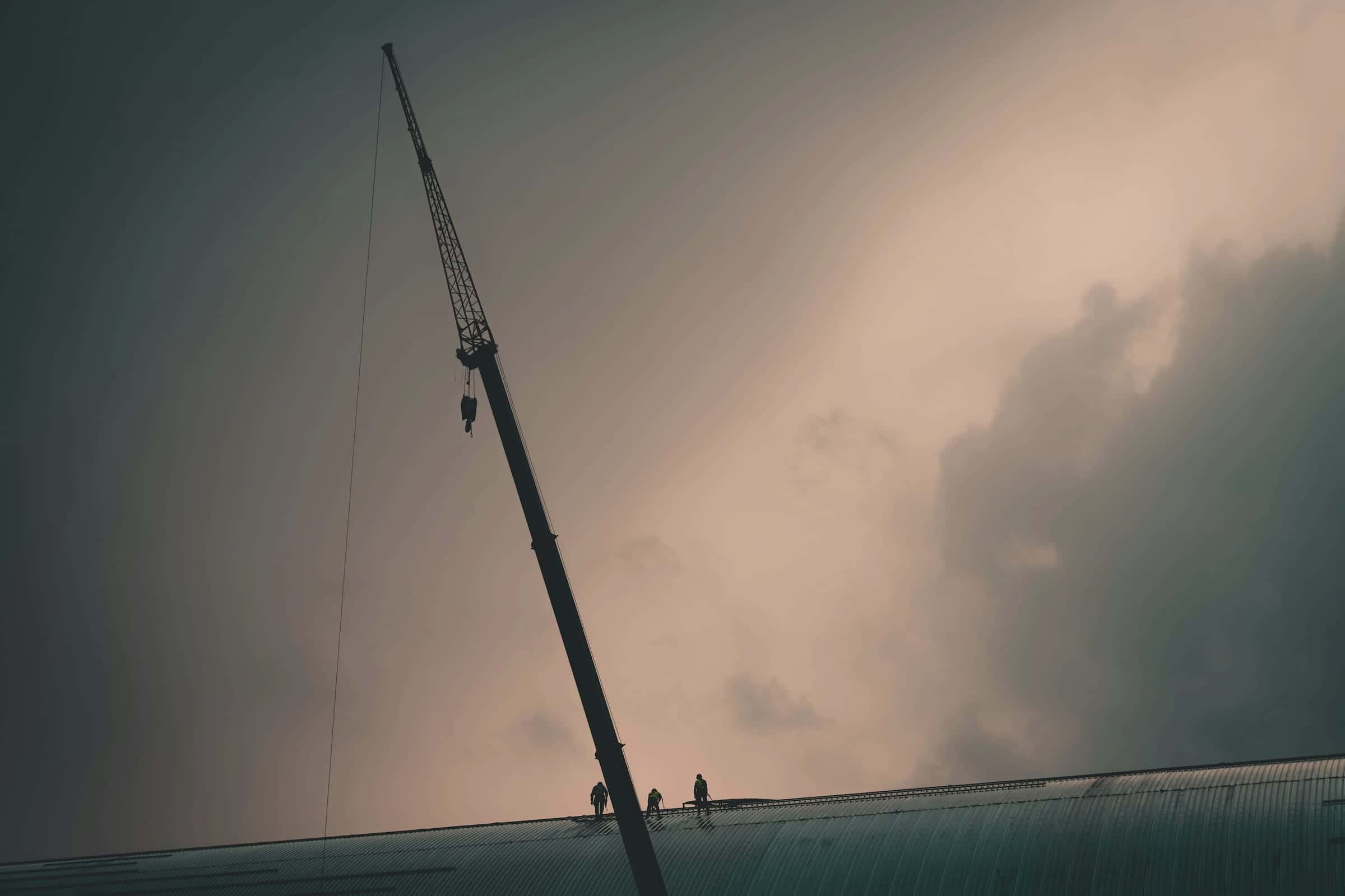 Three workers in hard hats and harnesses on a curved roof beside a crane, against a cloudy sky.