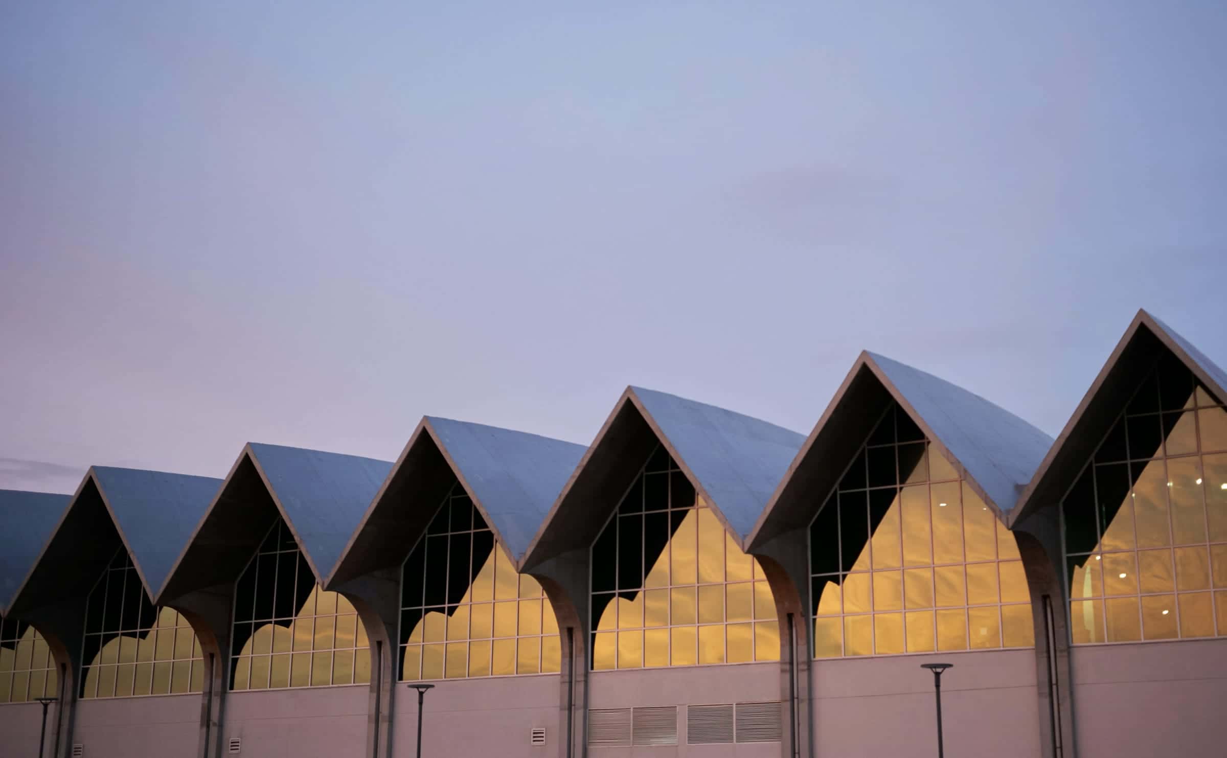 A row of pointed shell roofs over lit glass halls against a violet evening sky.