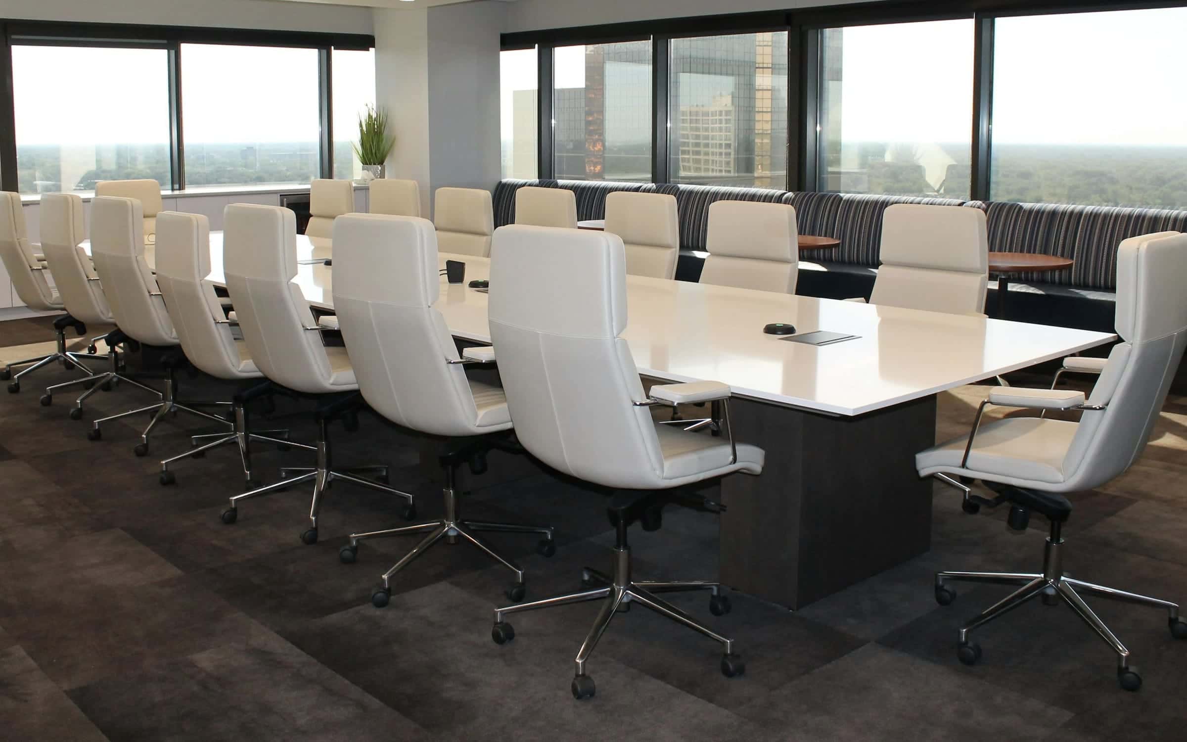 A long white meeting table with white chairs, beside windows over a wooded city.