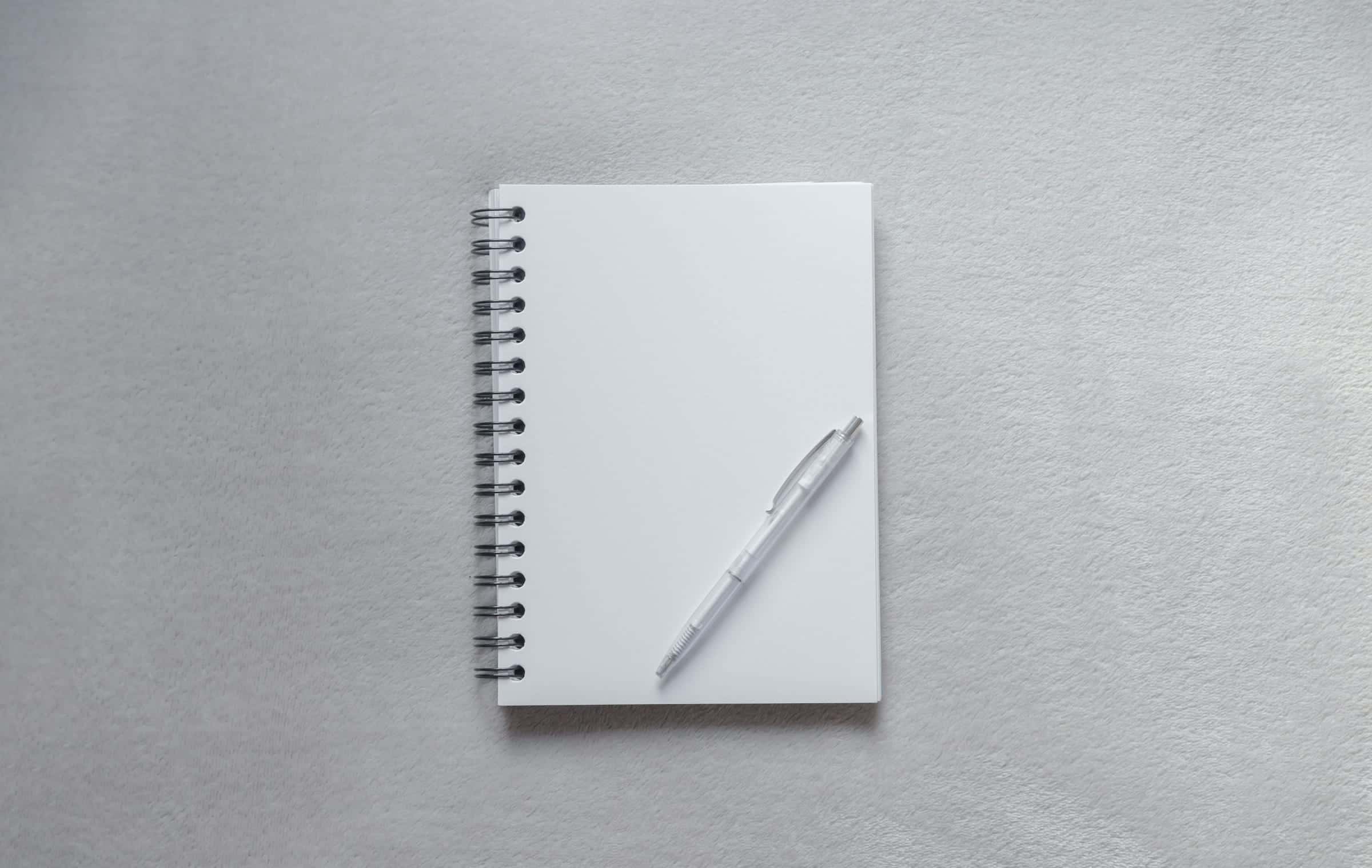 A closed white notebook and a pen on a pale grey surface.