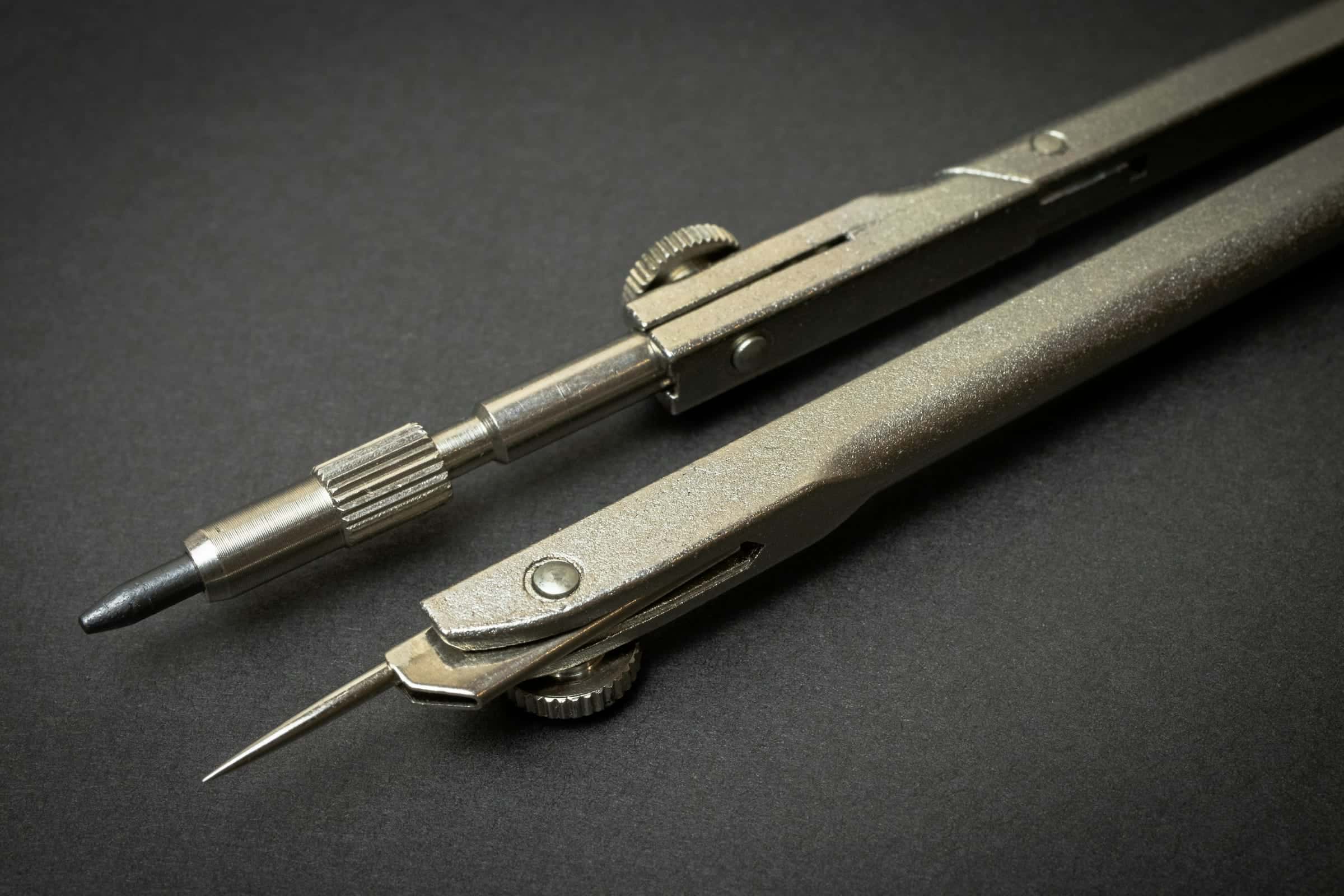 A drafting compass with a lead point and a needle point, resting on a dark surface.