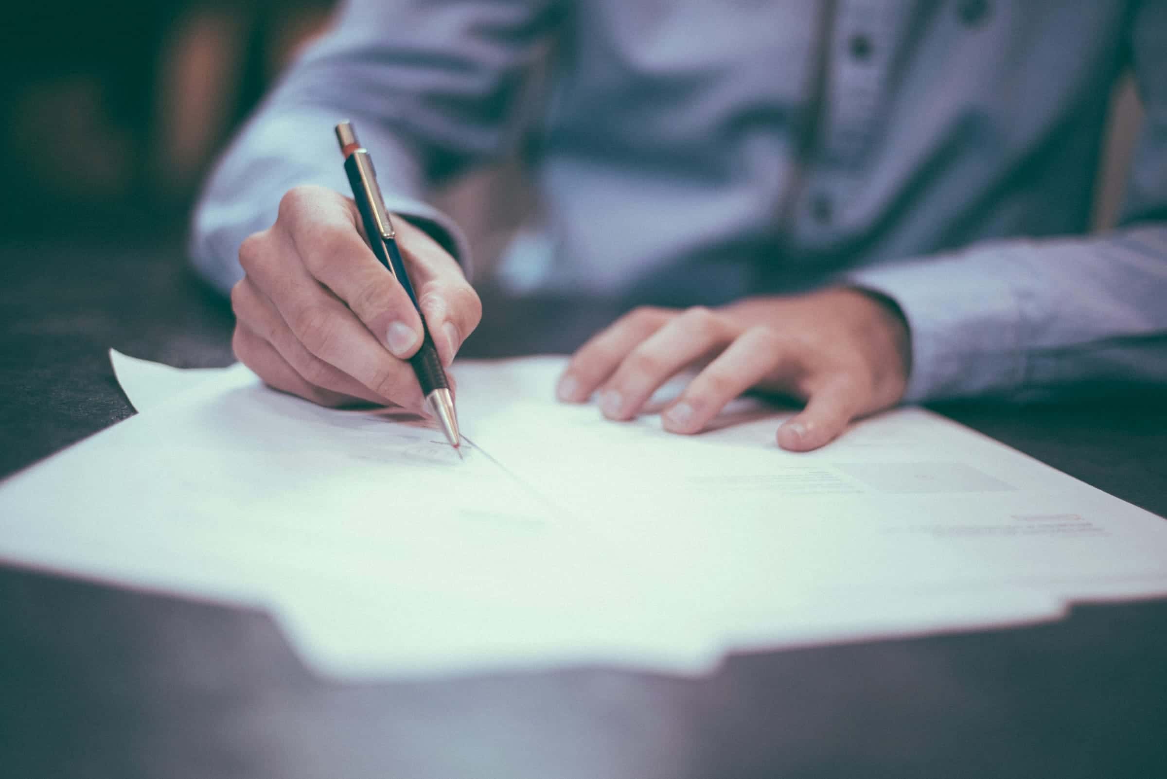 A hand signing a document with a pen on a dark table.