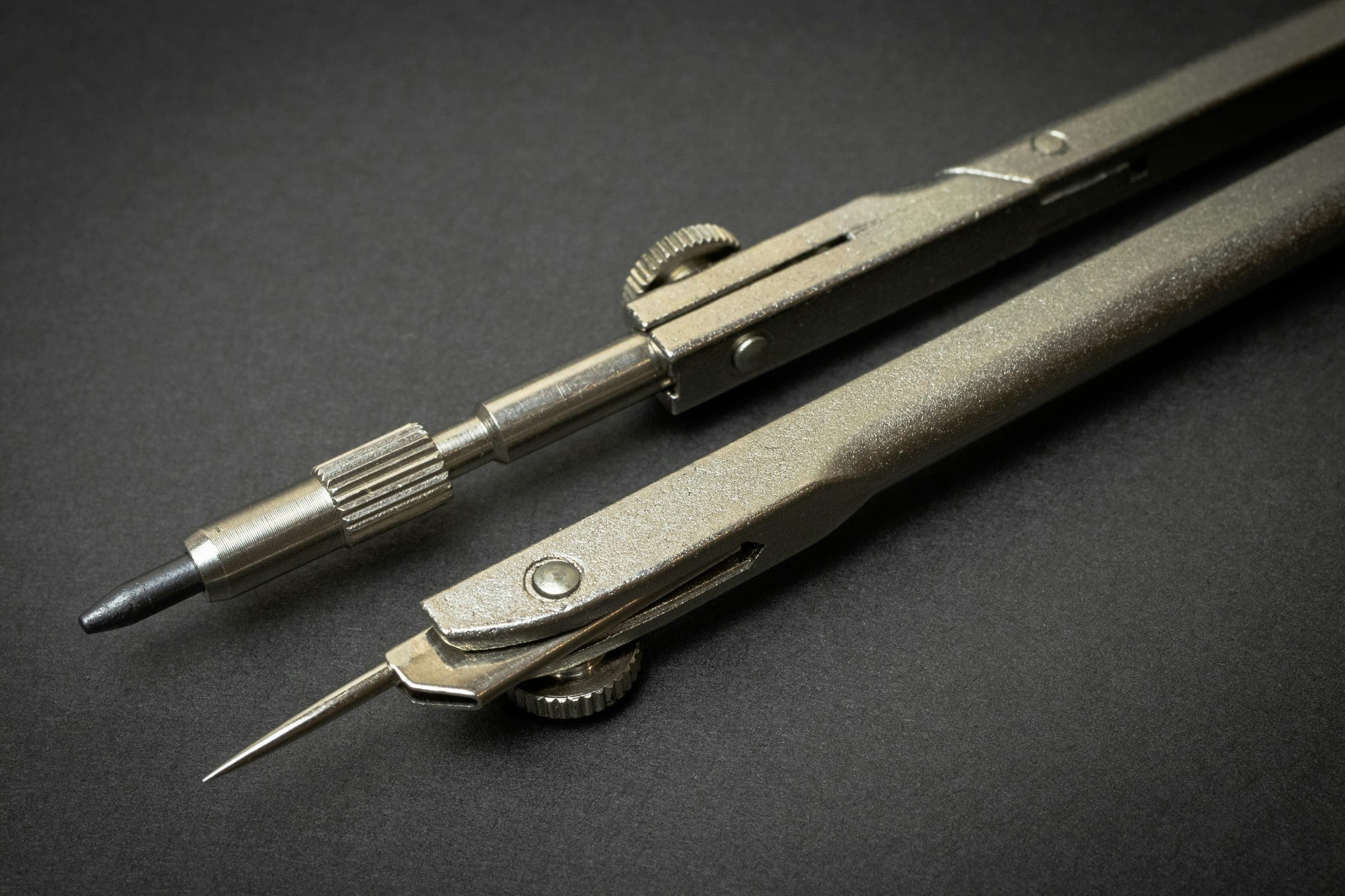 A drafting compass with a lead point and a needle point, resting on a dark surface.